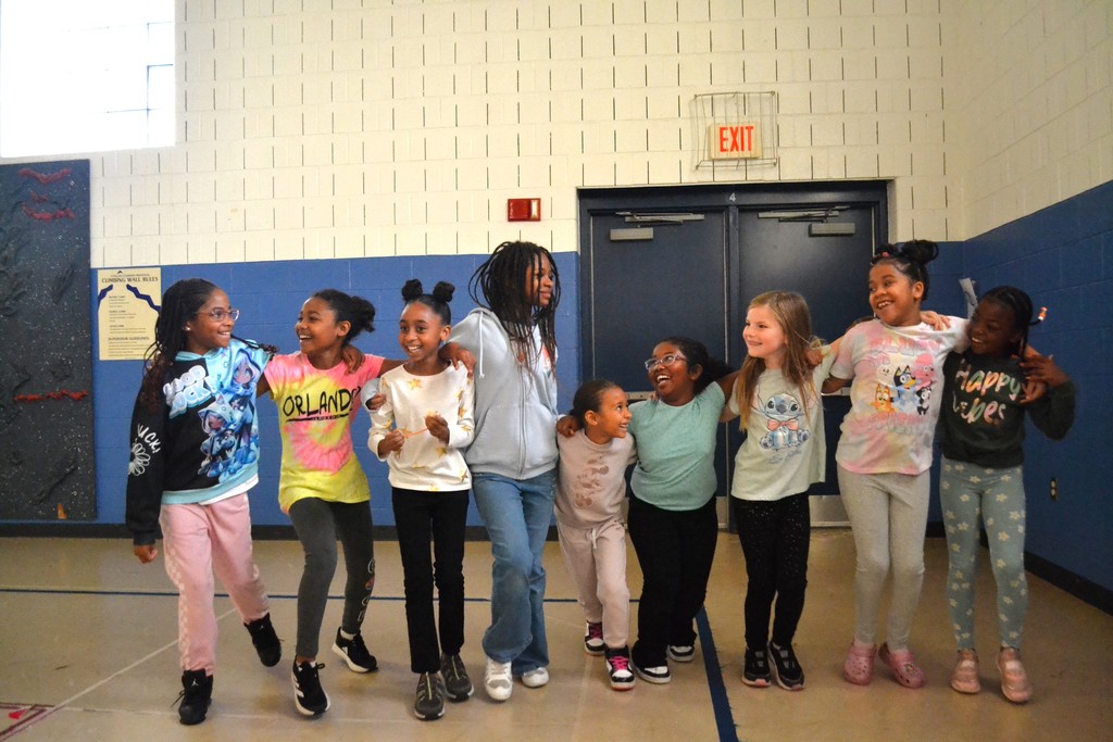 Image contain 9 girls smiling in a line linked arm and arm in the school gym.