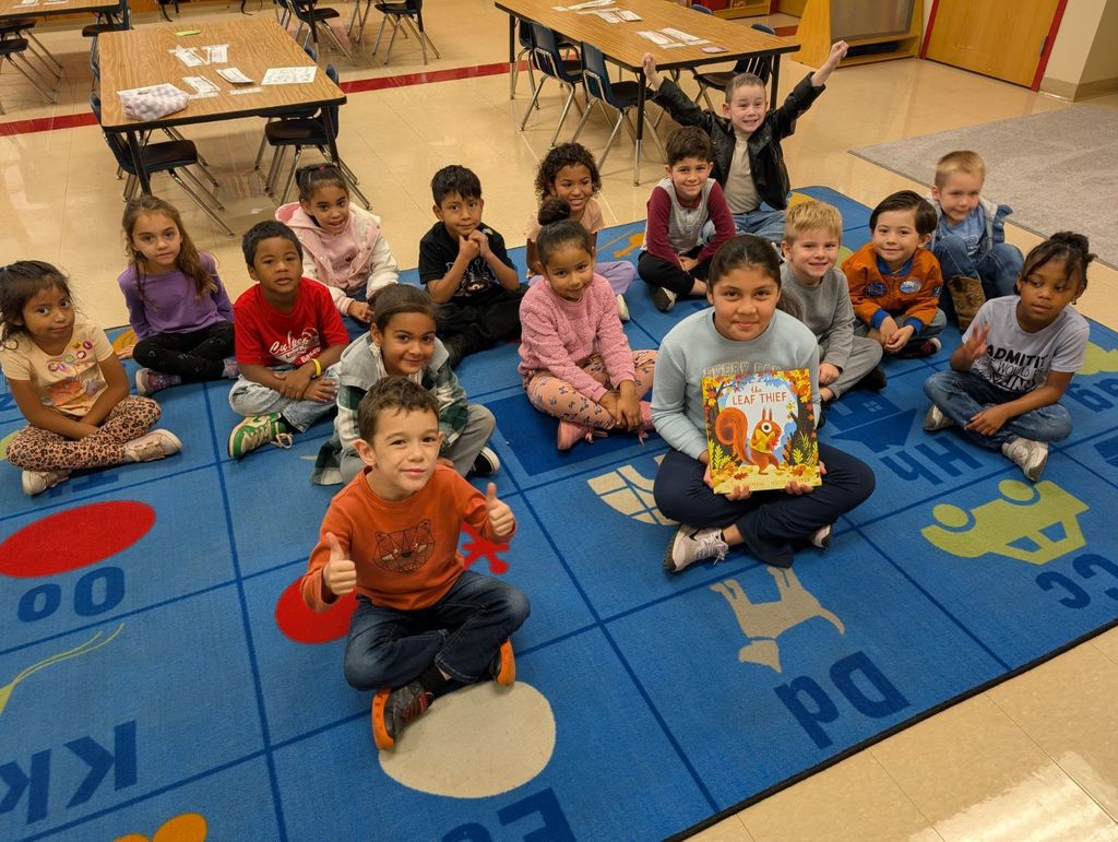 A group of young students sit smiling on a blue classroom carpet decorated with letters and pictures. In the center, a fifth-grade student named Allison holds up the book The Leaf Thief after reading it to the class. The children around her are seated cross-legged, some giving thumbs up, and everyone looks happy and engaged.