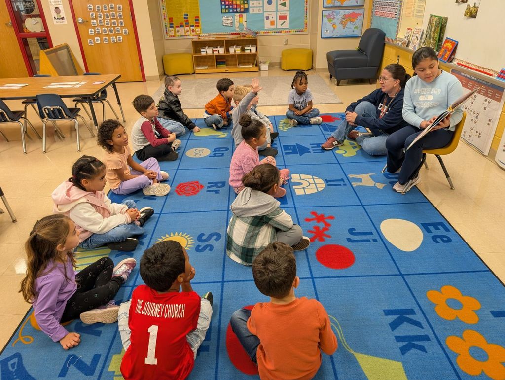 Allison, a fifth-grade ELL student, sits in a yellow chair reading The Leaf Thief aloud to a younger class seated on a colorful alphabet carpet. The younger students listen attentively, some raising their hands, while the teacher sits beside Allison smiling and supporting the activity.