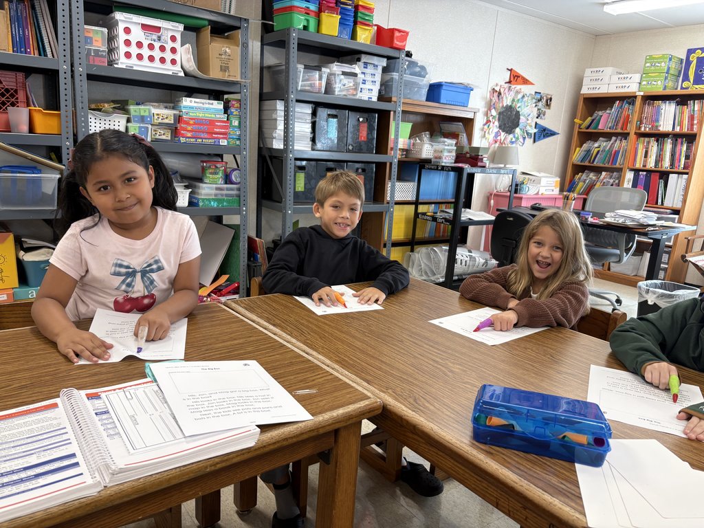 A group of four smiling elementary school-aged children seated at wooden desks in a classroom. The girl on the left is looking directly at the camera, as is the girl on the right. Shelves filled with colorful supplies and books are visible in the background. They are all working on assignments.