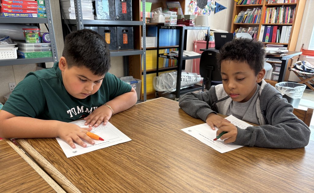 Two elementary school-aged boys sit at a wooden desk, focused on working on a worksheet that features text. The boy on the left wears a green "Tommy" shirt and is holding an orange highlighter, and the boy on the right wears a grey hooded sweatshirt and holds a green marker. Classroom shelves filled with books and supplies are in the background.