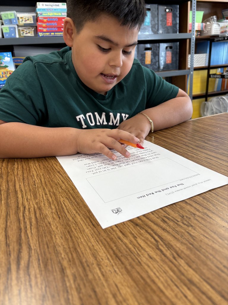 A close-up of an elementary school-aged boy wearing a green "Tommy" shirt, intensely focused on reading a worksheet titled "The Fox and the Red Hen." He is holding a pencil over the text.