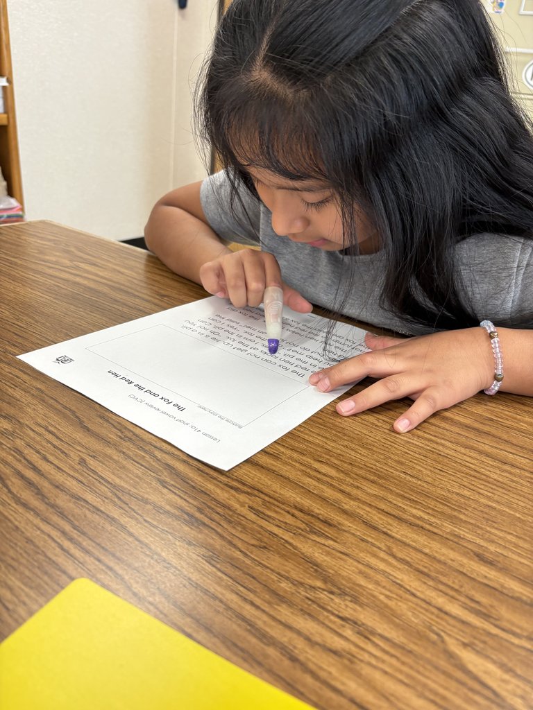 A close-up of an elementary school-aged girl with long dark hair, wearing a grey shirt and a beaded bracelet, focused on a worksheet titled "The Fox and the Red Hen." She is holding a purple marker or highlighter over the text.