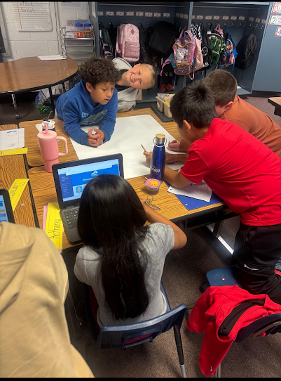  A group of five elementary school students collaborating on a project in a classroom. They are gathered around a table with a large piece of white paper and an open laptop. One girl is seated, looking at the screen, while four other students lean over the paper to write or draw.