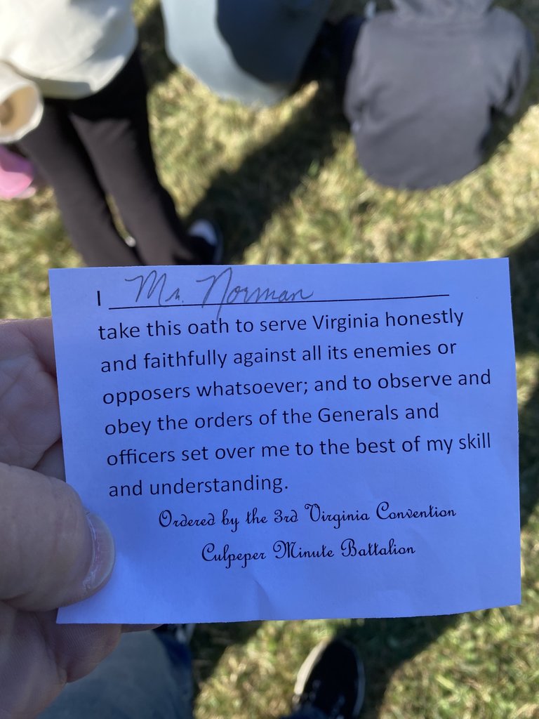A hand holds a small piece of paper with a handwritten name "Mr. Norman" and a printed oath below: "take this oath to serve Virginia honestly and faithfully against all its enemies or opposers whatsoever; and to observe and obey the orders of the Generals and officers set over me to the best of my skill and understanding. Ordered by the 3rd Virginia Convention Culpeper Minute Battalion."