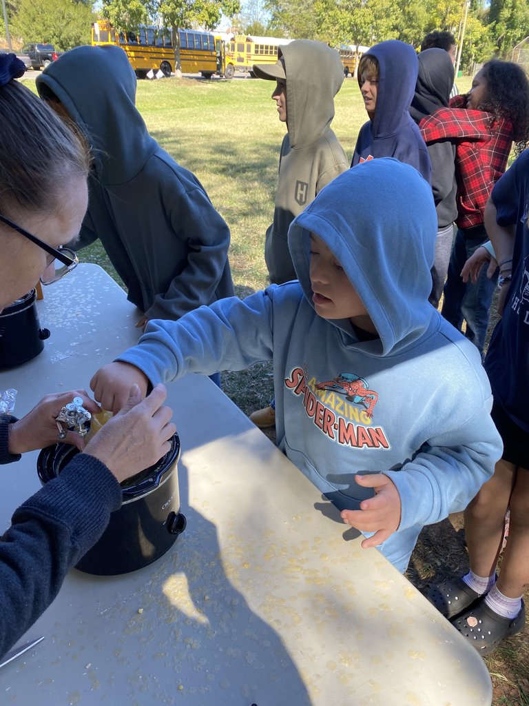 A young boy in a blue Spider-Man hoodie is being helped by an adult to handle a piece of aluminum foil over a small black slow cooker on a table, likely participating in a historical activity or demonstration.