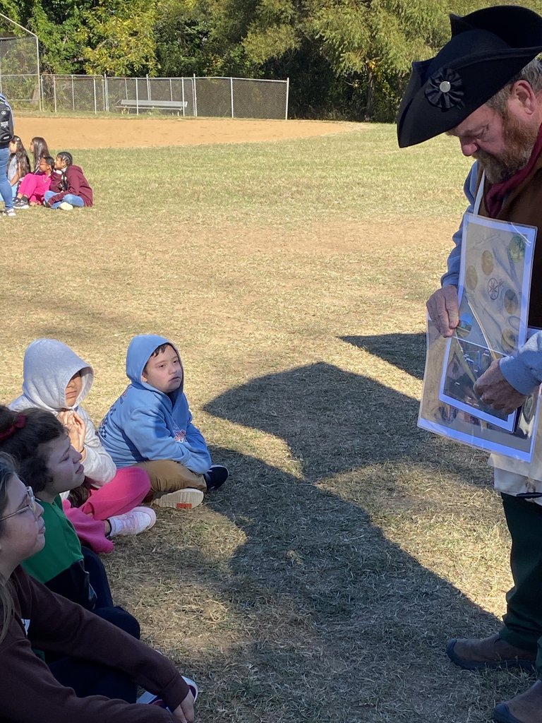 A young boy in a blue Spider-Man hoodie is being helped by an adult to handle a piece of aluminum foil over a small black slow cooker on a table, likely participating in a historical activity or demonstration.