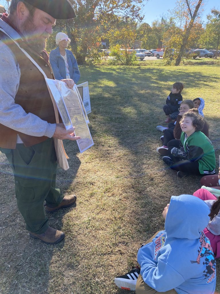 A historical reenactor in a brown vest and tricorn hat, holding laminated pictures or maps, speaks to a group of children sitting on the grass.