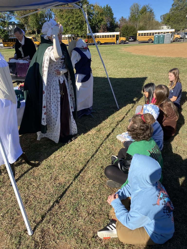 A reenactor, partially visible, helps a young girl in a green and black varsity jacket handle a small, round object over a heating element on a table, possibly demonstrating a historical cooking or craft method. IMG_9899.jpg: A historical reenactor in a tricorn hat and brown vest is speaking to a group of children sitting on the grass, holding up laminated pictures to illustrate his talk. IMG_9894.jpg: A female historical reenactor in a long white dress and a dark green cloak speaks to a group of children sitting on the grass. Other reenactors and tents are visible in the background.
