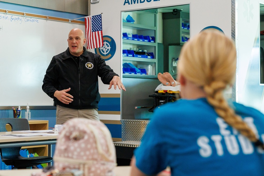 A law enforcement officer presents to EMT students in a CTEC classroom equipped with an ambulance training simulator in the background.