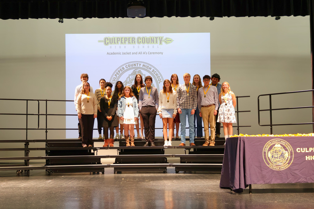 A large group of students stands on risers on a stage wearing medals around their necks during the Culpeper County High School Academic Jacket and All A’s Ceremony. A presentation slide with the school logo is projected behind them.