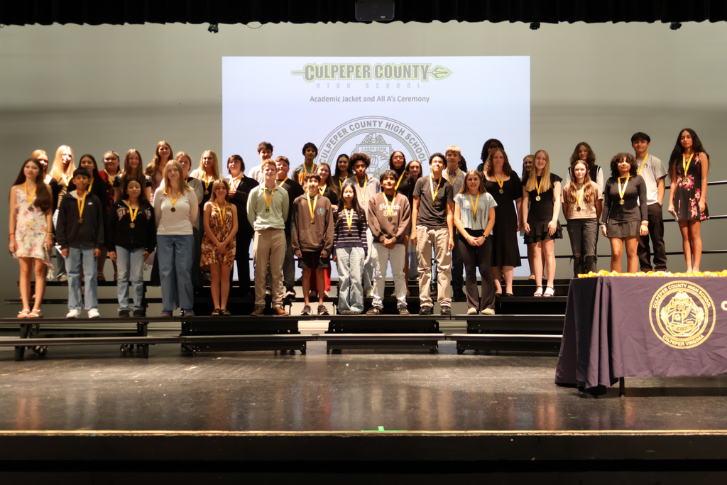 A large group of students stands on risers on a stage wearing medals around their necks during the Culpeper County High School Academic Jacket and All A’s Ceremony. A presentation slide with the school logo is projected behind them.