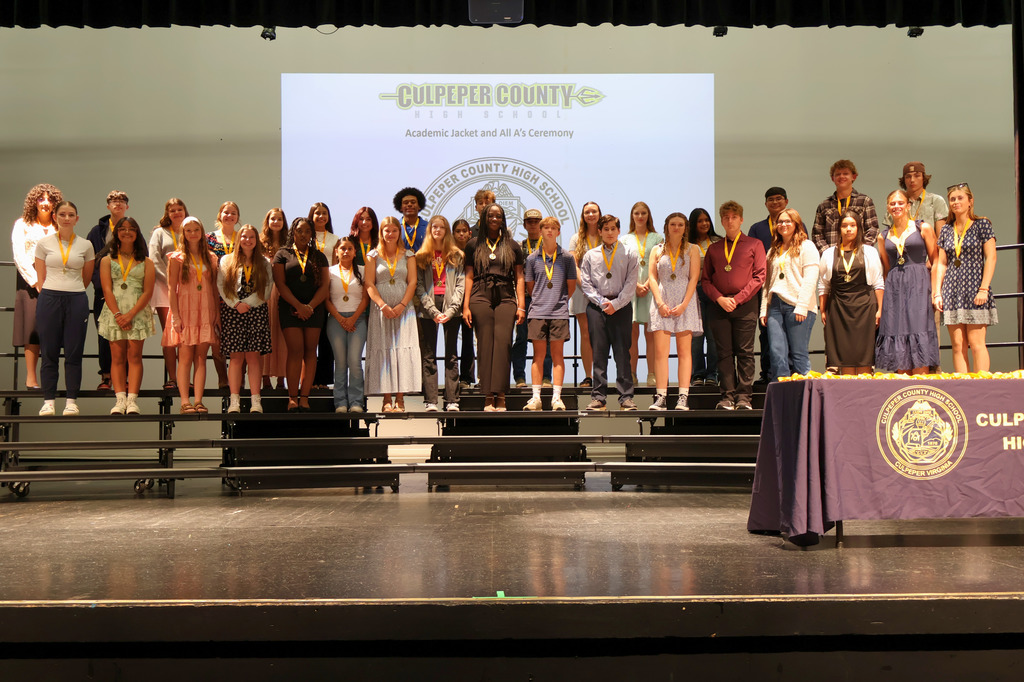 A large group of students stands on risers on a stage wearing medals around their necks during the Culpeper County High School Academic Jacket and All A’s Ceremony. A presentation slide with the school logo is projected behind them.