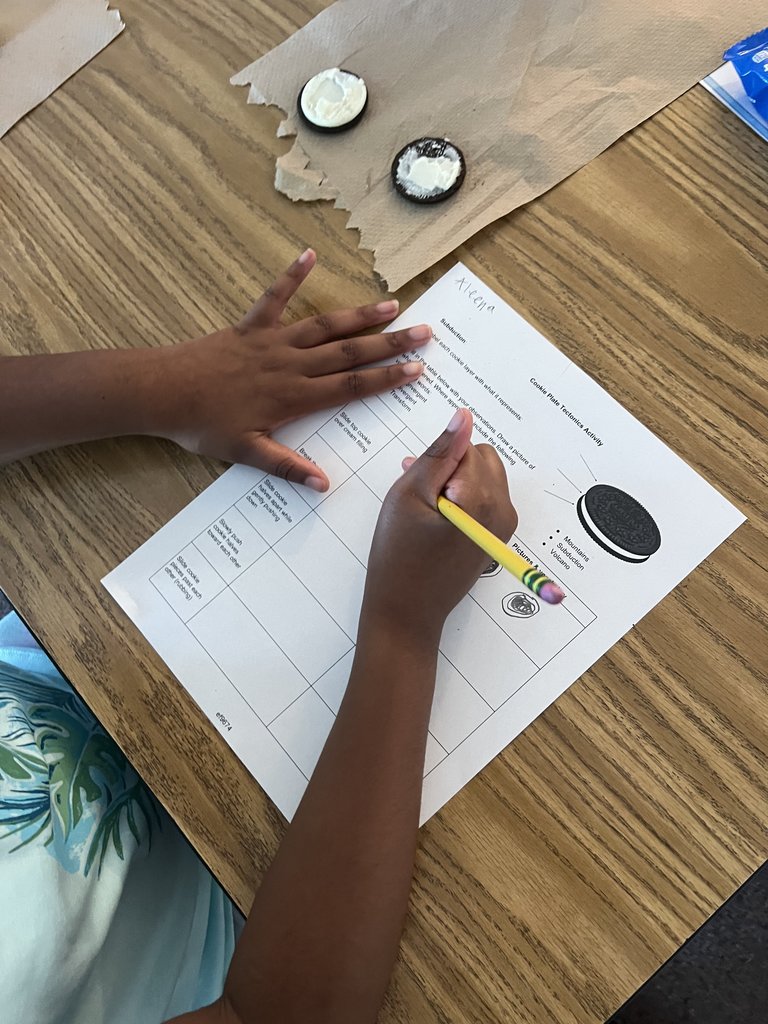 A close-up shot of a student's hands writing on a worksheet titled "Cookie Plate Tectonics Activity" with a yellow pencil. Two cream-filled cookie halves are resting on a brown paper towel on a wooden desk. The worksheet outlines different types of plate boundaries: convergent, divergent, and transform, using the cookie parts as models.