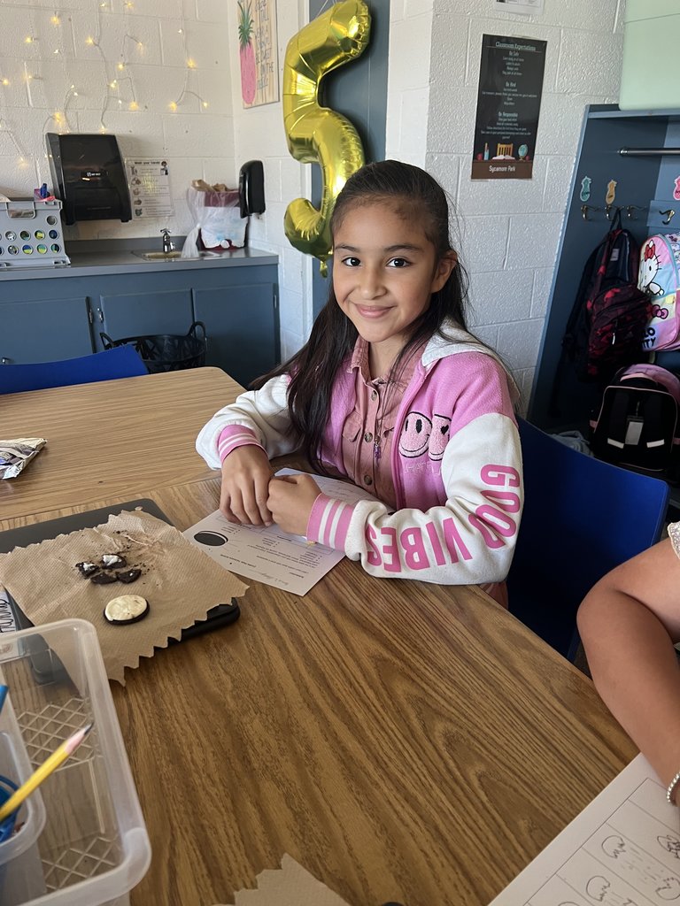A young girl with long, dark hair wearing a pink and white sweatshirt with a smiling face graphic is seated at a table, looking up and smiling at the camera. A partially eaten cookie is on a paper towel on the table in front of her. The classroom background includes colorful decorations and signs.
