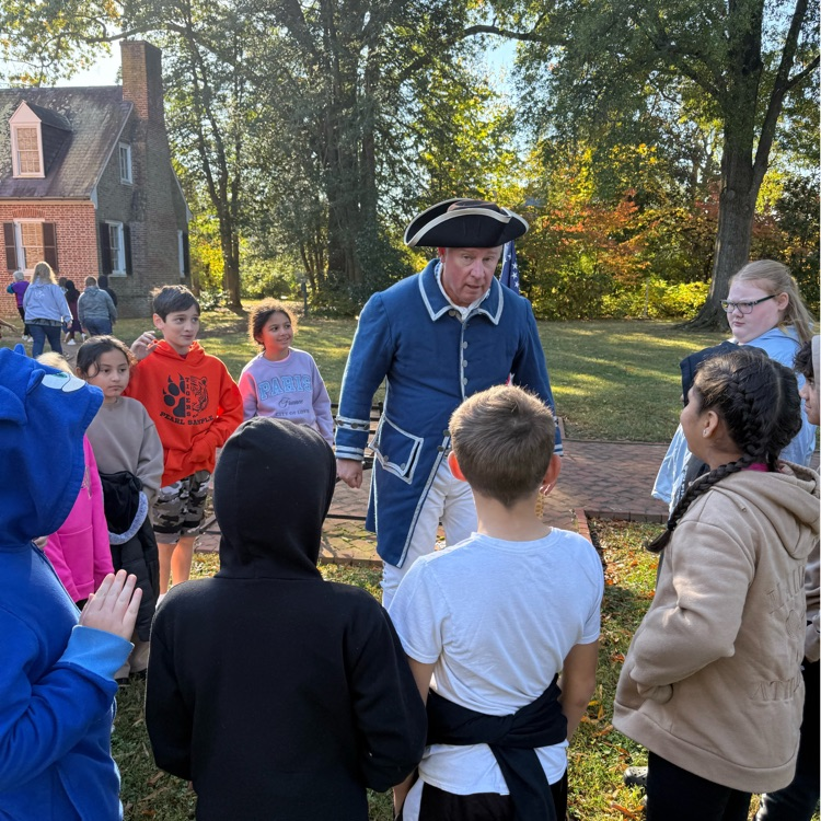 A group of students and an adult in colonial era clothing stand outside a brick building on a sunny day