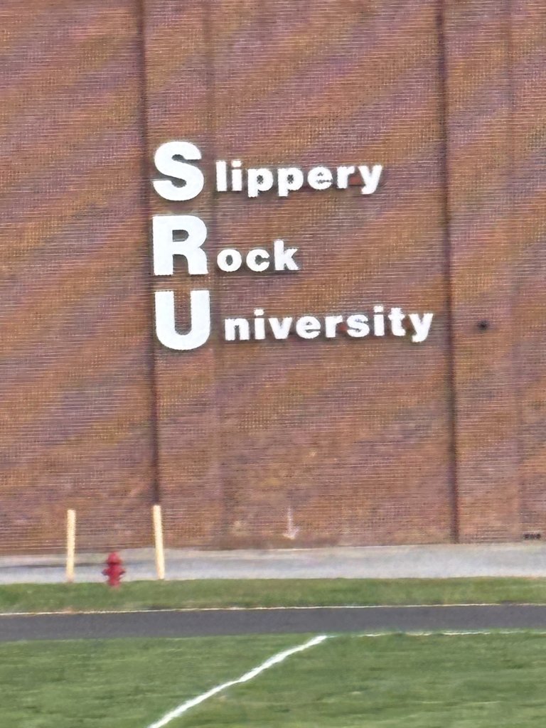 A close-up, cropped view of a brick building with large white letters spelling out "SRU Slippery Rock University". A fire hydrant and the edge of a grassy field are visible below.