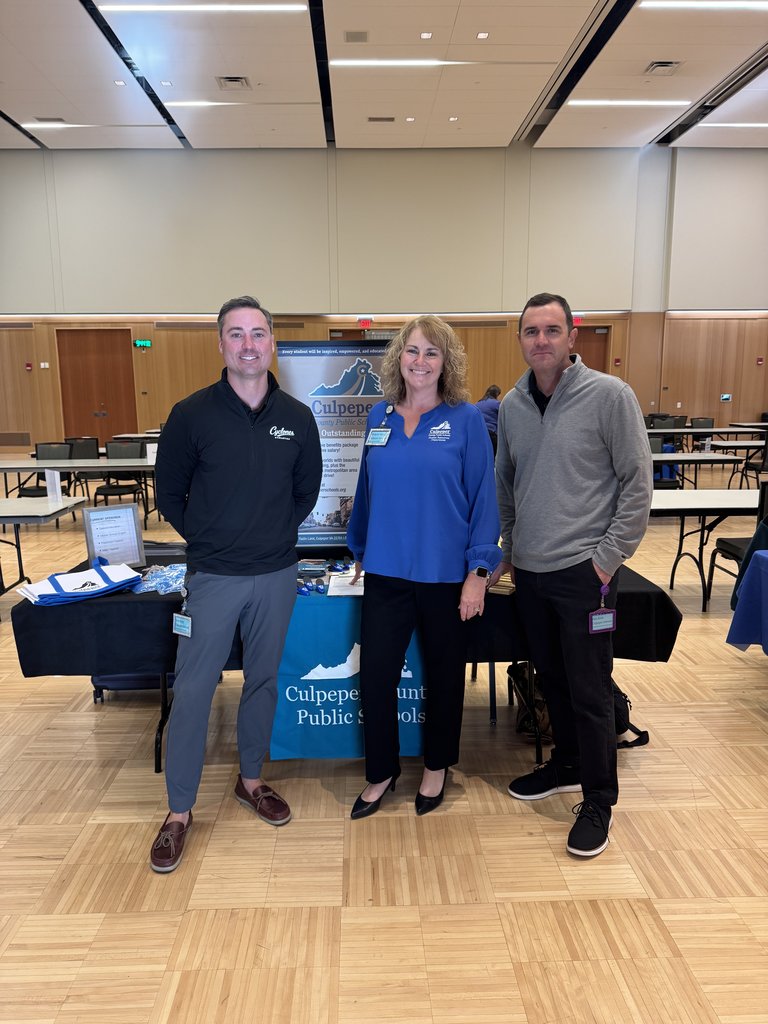Three adults (two men and one woman) standing behind a table with a banner for "Culpeper County Public Schools" at an indoor event or convention.