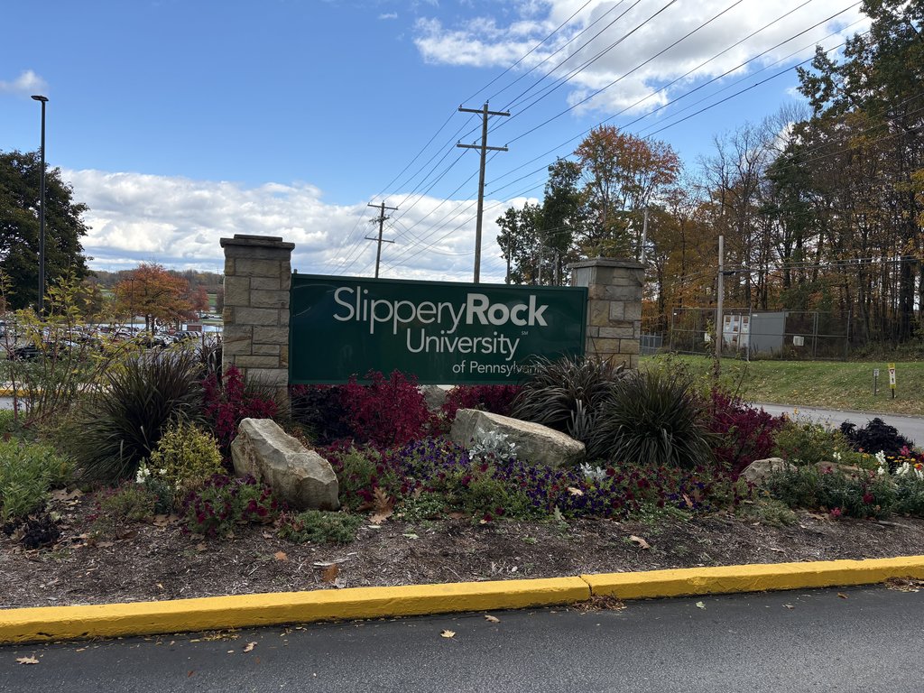 A stone and green sign for "Slippery Rock University of Pennsylvania" surrounded by landscaping and rocks, on a sunny day with clouds.