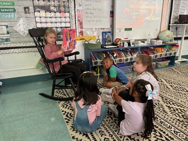A young girl with blonde hair, wearing a pink top, sits in a dark wooden rocking chair reading a book out loud to a small group of four other students. The listeners are sitting attentively on the patterned black and white rug in front of her. The background shows a cozy classroom corner with a colorful book display on shelves, decorations, and a whiteboard listing classroom rewards.