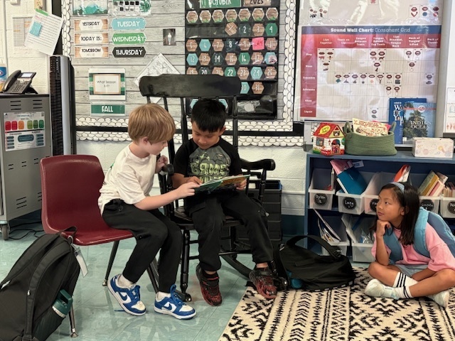 Two young boys are sitting side-by-side in a reading corner. One boy, wearing a white shirt, is seated on a red chair while the other, wearing a black shirt, is in a wooden rocking chair. They are both looking down at a picture book, sharing a moment of reading. Another girl sits on the floor to the right, looking up at them. Classroom decorations, a wall calendar, and a sound chart are visible in the background.