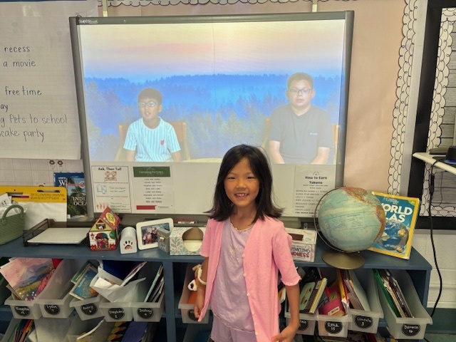 A young girl with a bright smile, wearing a pink cardigan, stands in front of a white screen displaying a muted blue and green image of two boys sitting in chairs, possibly from a video or presentation. She is standing behind a low shelf filled with books, a globe, and other classroom materials. The screen has a sign underneath that says "Ask Then Tell."