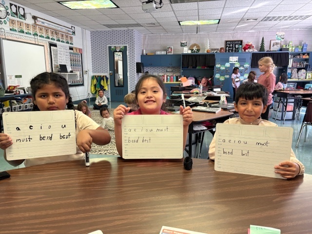 Three elementary-aged students are sitting at a dark brown table, proudly holding up individual whiteboards. Each whiteboard has handwritten letters and words: "a e i o u" on the top line, and a combination of words like "must bend best" written below. The students are looking directly at the camera, with a classroom setting visible behind them, including a rug, other students, and desks.