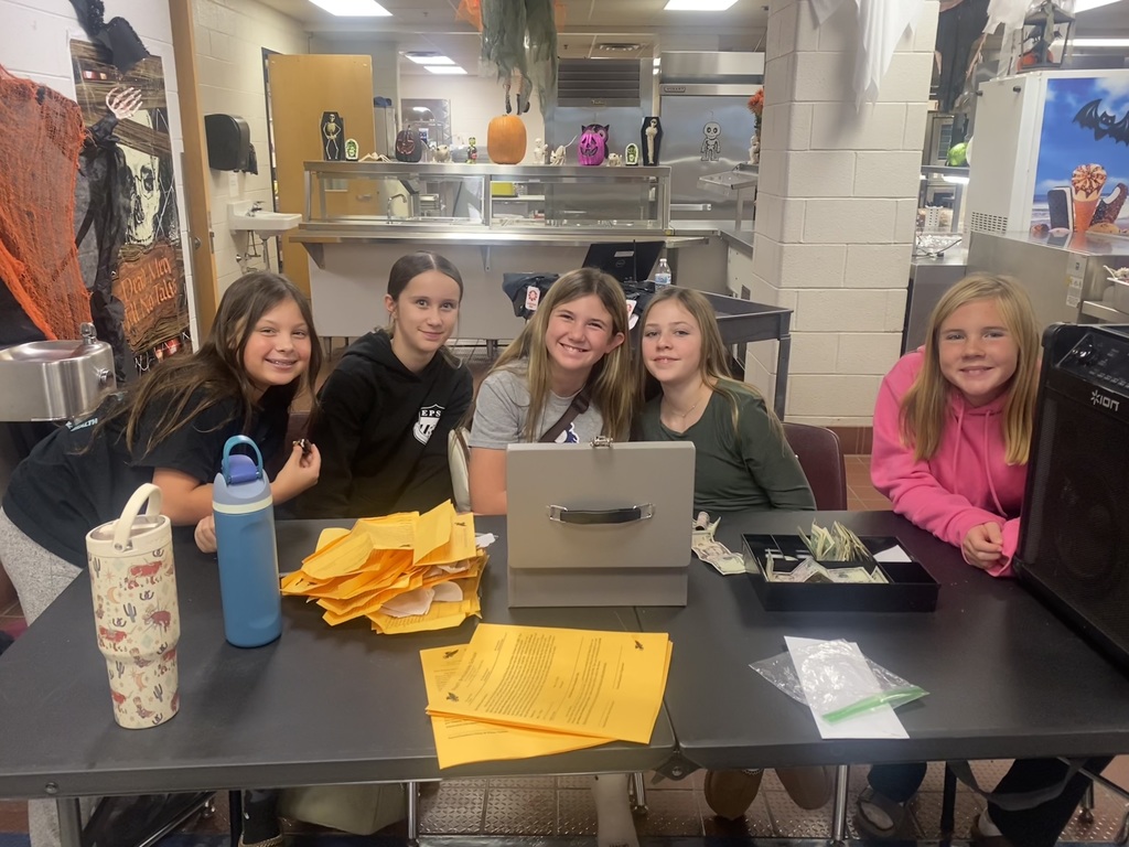 Five smiling girls sit at a table with stacks of papers and money, in a room decorated with Halloween-themed items like pumpkins and cobwebs.