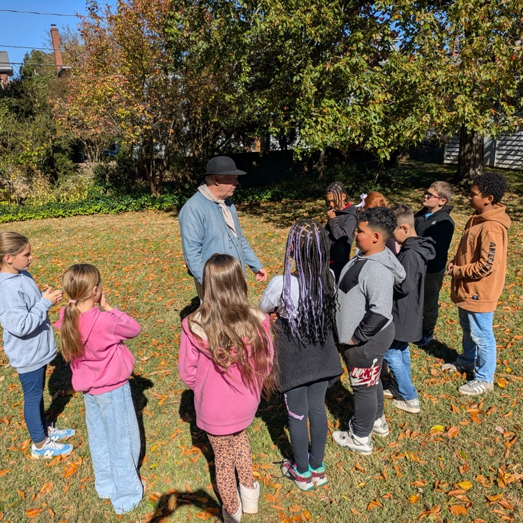 A group of students standing outside on a sunny day, listening to a man talk about life long ago, while dressed in colonial clothes