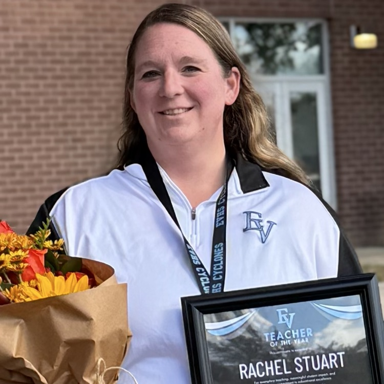 The image shows a woman standing outdoors in front of a brick building with fall-colored leaves around her. She is smiling and holding a framed award plaque in one hand and a bouquet of flowers wrapped in brown paper in the other.