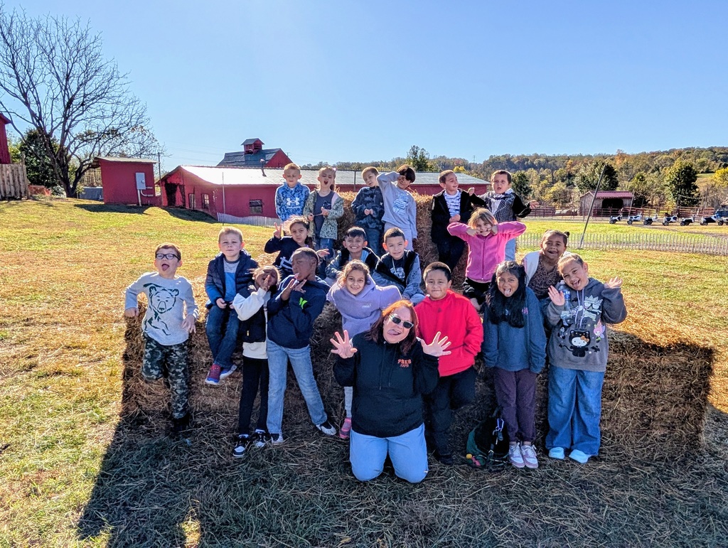A group of elementary school children and one adult female teacher are making silly faces and striking fun poses while standing and sitting on large hay bales in a sunny, grassy field. They are all smiling and enjoying the outdoor setting, with red farm buildings in the background.