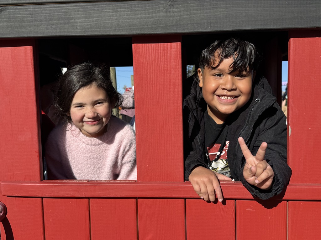 A close-up shot of two smiling children, a girl with dark hair wearing a light pink sweater and a boy with dark hair wearing a black jacket, looking out of the red wooden window of a small structure, possibly a train car or playhouse. The boy is holding up a peace sign.