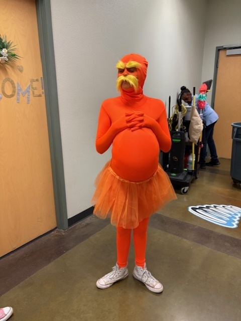A person wears an orange bodysuit with a matching tutu, yellow fluffy eyebrows, and mustache, resembling a playful character in a hallway setting.
