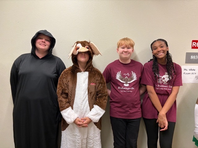 Four people stand against a wall. Two wear maroon T-shirts with eagle graphics, two are in costumes—a black robe and a furry animal outfit. They appear relaxed and cheerful.