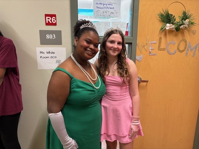 Two women smiling in colorful dresses stand in front of a decorated classroom door with a "Welcome" sign. They wear tiaras and long gloves.