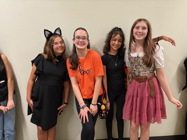 Four girls in festive attire stand smiling against a light wall. They wear costumes, including cat ears and a folk dress, conveying a joyful and playful mood.