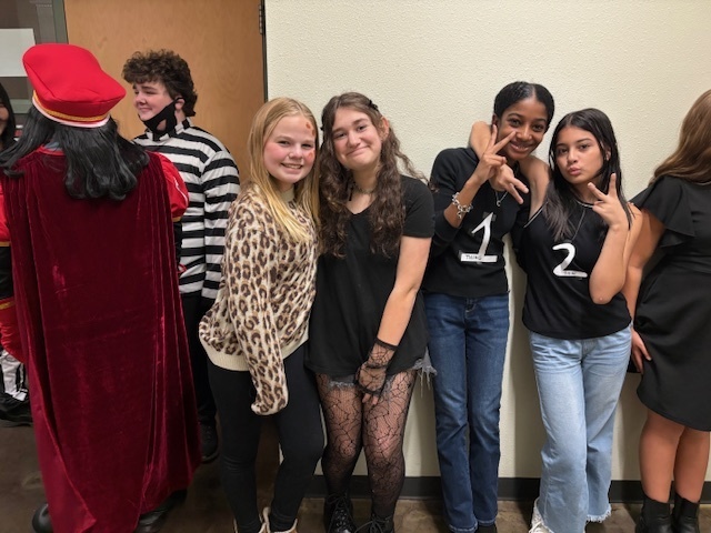 A group of four smiling girls poses together in stylish outfits. Behind them, a person in a red costume and another in black and white stripes are visible. The scene conveys a fun and friendly atmosphere.