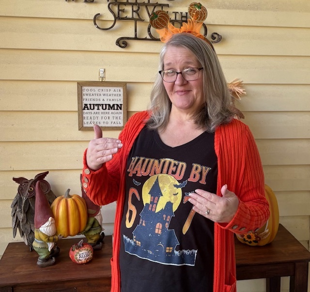 A woman in a festive shirt and orange cardigan smiles, pointing at her "Haunted by 6 7" design. Pumpkins and a gnome decorate the autumn-themed scene.