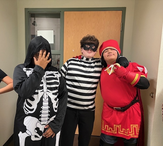 Three people in costumes stand in front of a door, smiling. One wears a skeleton outfit, another is dressed as a thief, and the third is in a medieval knight costume.