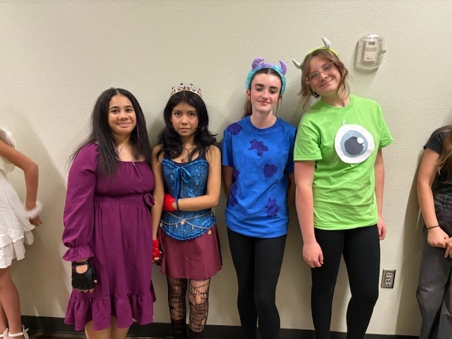 Four individuals stand against a light wall in costumes. They appear joyful and playful, showcasing various colorful outfits and accessories.