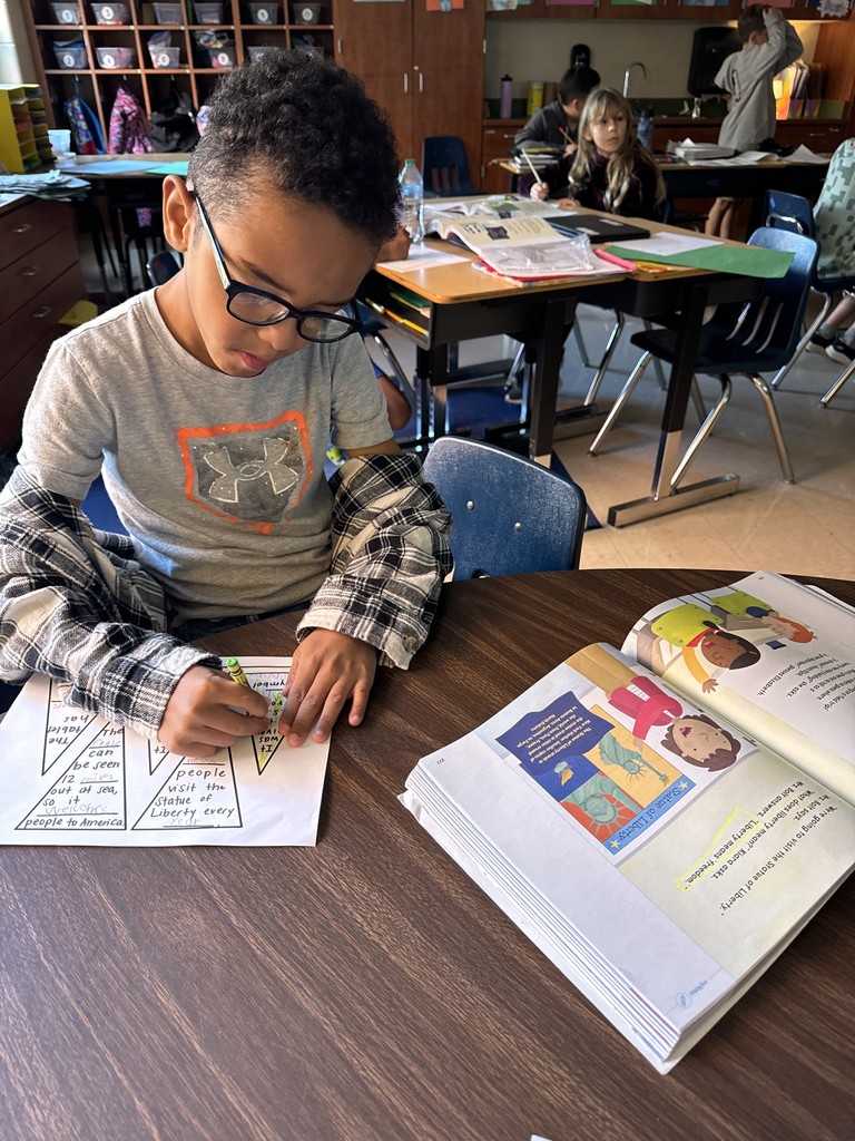 A student wearing glasses colors a green Statue of Liberty crown worksheet while referencing a reading book open on the desk beside him.