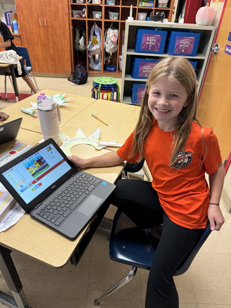 . A student in an orange shirt smiles at her desk while showing her Chromebook screen featuring a Scratch project with a colorful city background.