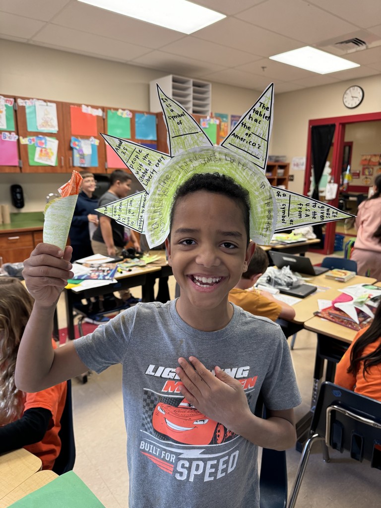 A smiling student wearing a gray shirt with Lightning McQueen on it holds a handmade paper torch and wears a green paper Statue of Liberty crown with facts written on the spikes.