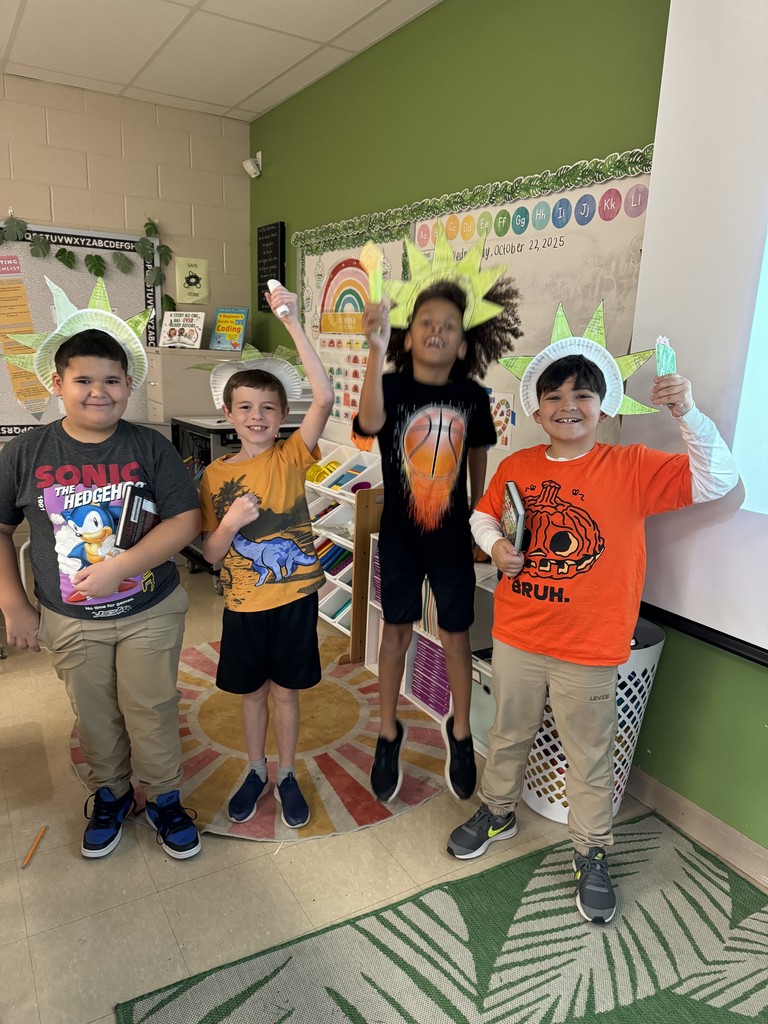 Four students pose in front of a classroom rainbow bulletin board, three wearing Statue of Liberty crowns and holding paper torches, while one jumps midair excitedly.