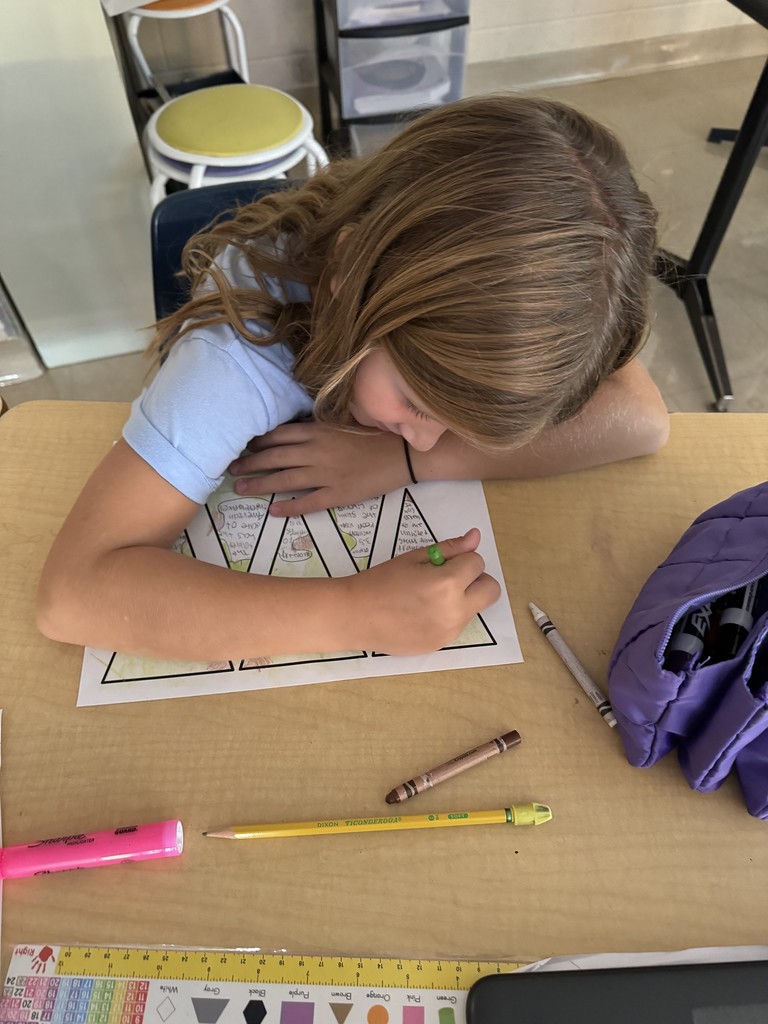 A student colors a Statue of Liberty crown worksheet with green crayon at her desk, surrounded by school supplies including a pink highlighter, pencil, and purple pencil case.