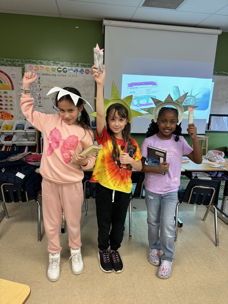 Three students pose proudly in front of a classroom screen, wearing Statue of Liberty crowns and holding paper torches. One wears a pink outfit, one a tie-dye shirt, and one a lavender shirt with jeans.