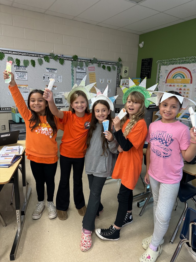 Five students stand in a classroom smiling and holding paper torches while wearing handmade Statue of Liberty crowns. The bulletin board behind them displays classroom reading and writing materials.