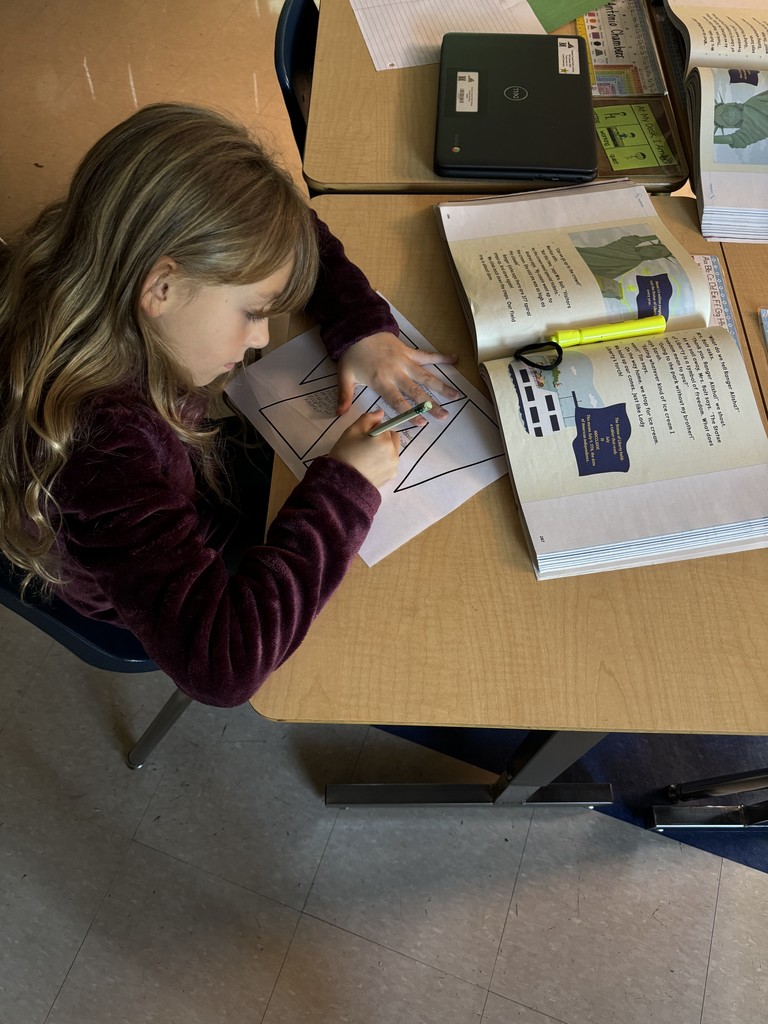 A student with long blond hair works at her desk coloring a triangular worksheet with facts about the Statue of Liberty, using a pencil and green crayon.