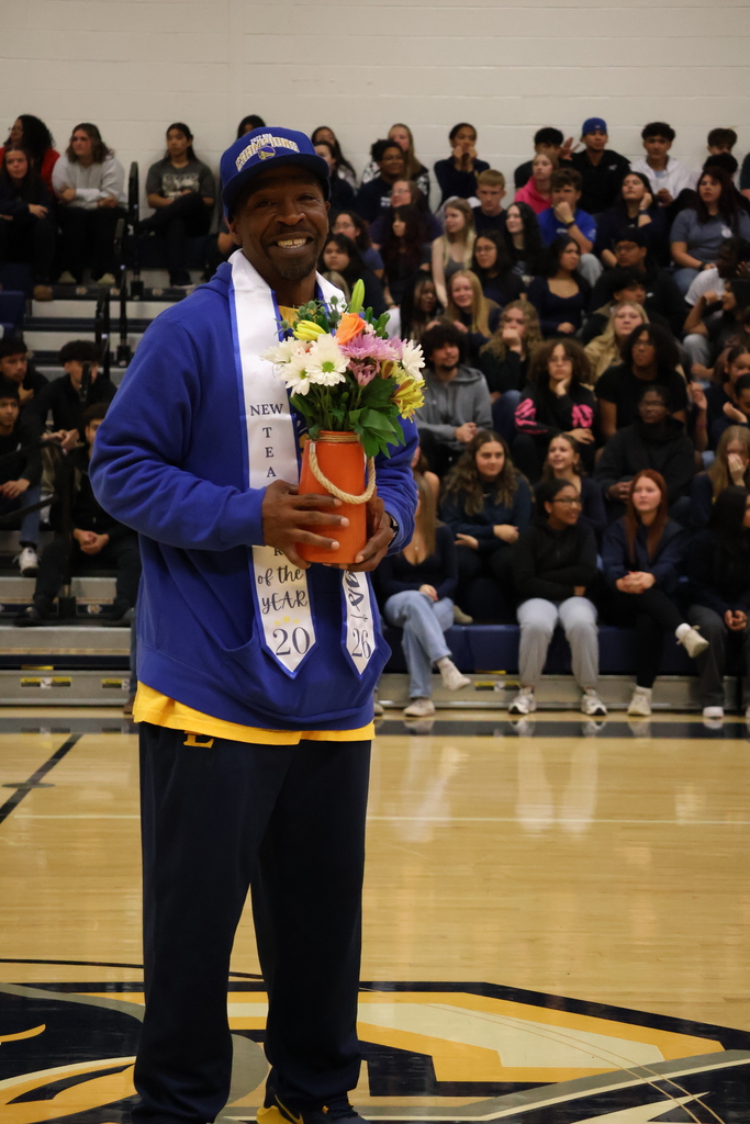 A smiling teacher stands on a gym floor holding a bouquet of flowers in an orange vase. He wears a blue and gold school sweatshirt, matching sweatpants, and a blue cap. A white sash around his neck reads “New Teacher of the Year 2026.” Students sit in the bleachers behind him, watching the event.