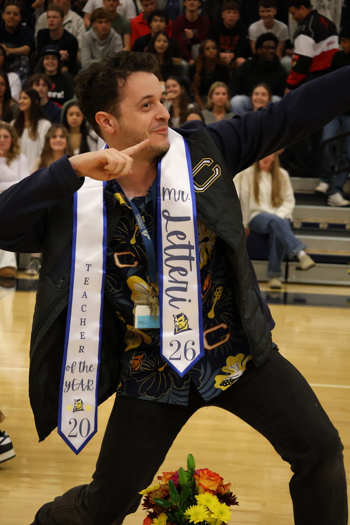 A teacher wearing a dark patterned shirt and jacket poses playfully on a gym floor, pointing upward in a celebratory stance. He wears two white sashes that read “Teacher of the Year 2026” and “Mr. Letteri.” A bouquet of colorful flowers rests on the floor in front of him, and a crowd of students sits in the bleachers behind him, watching and smiling.